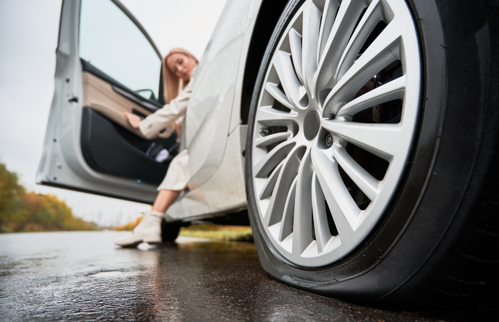 Female driver of white car observing punctured tire on rear wheel.