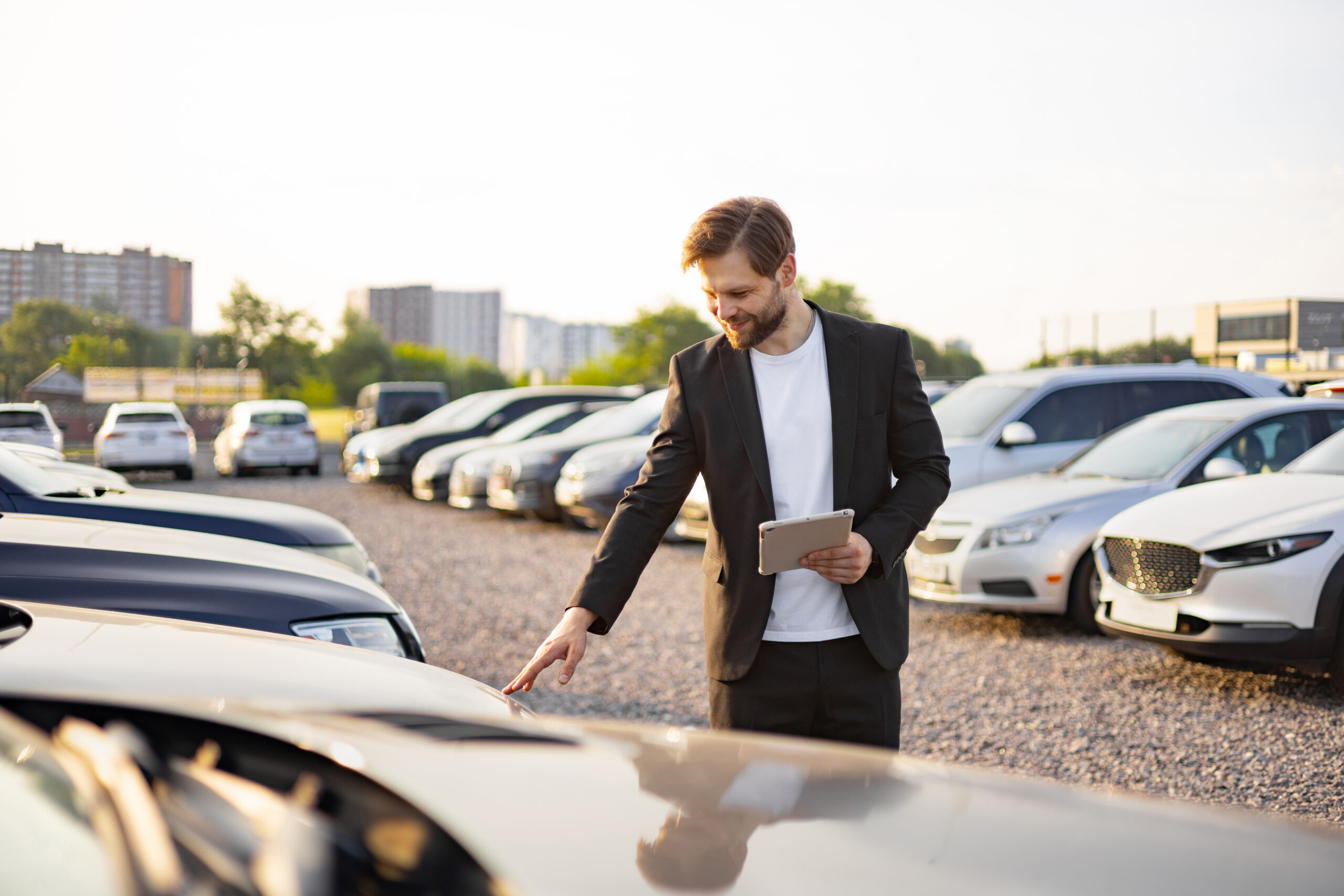 A car salesman checks a car at a dealership. He is holding a tablet while looking at the car with a smile, indicating a positive experience.