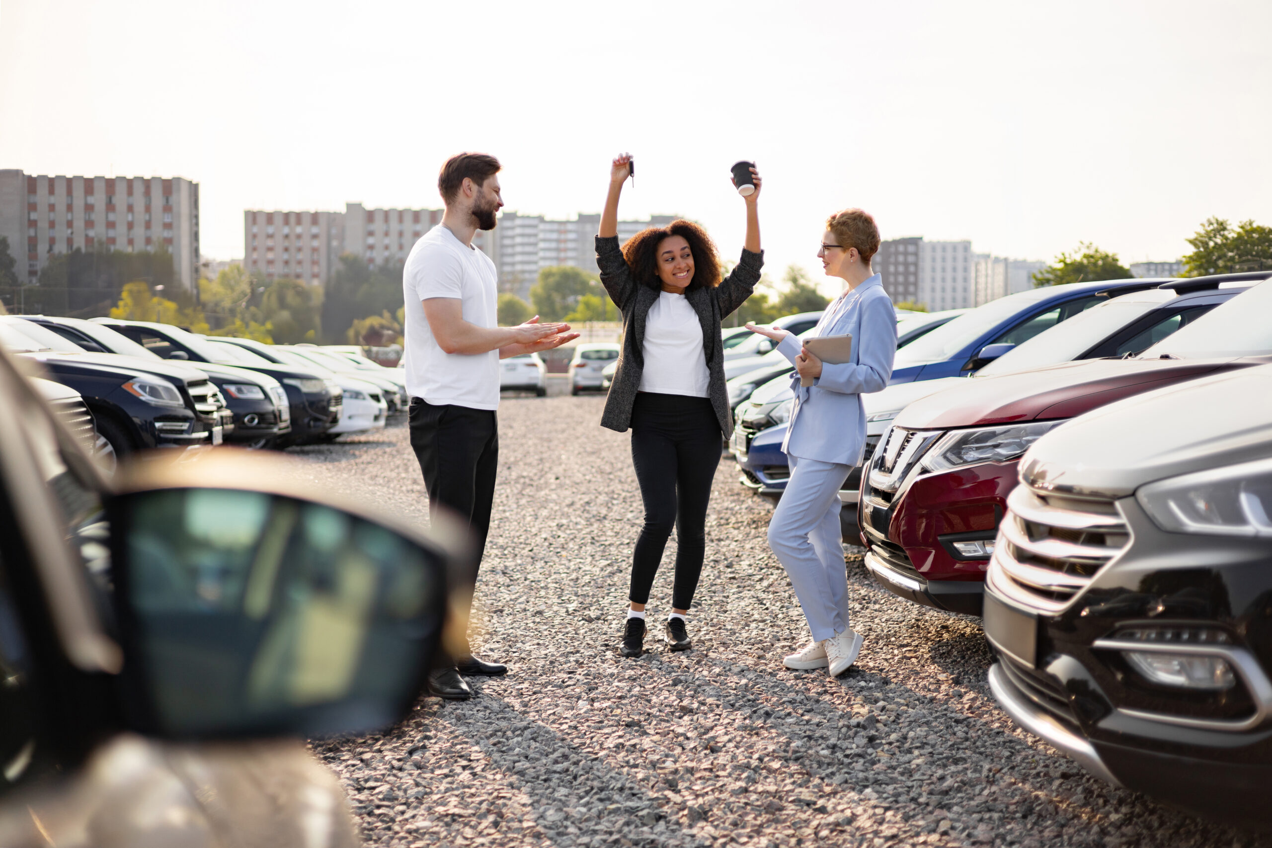 A woman celebrates her new car purchase at a dealership with a salesperson and her partner nearby. Cars of various models are in the background.
